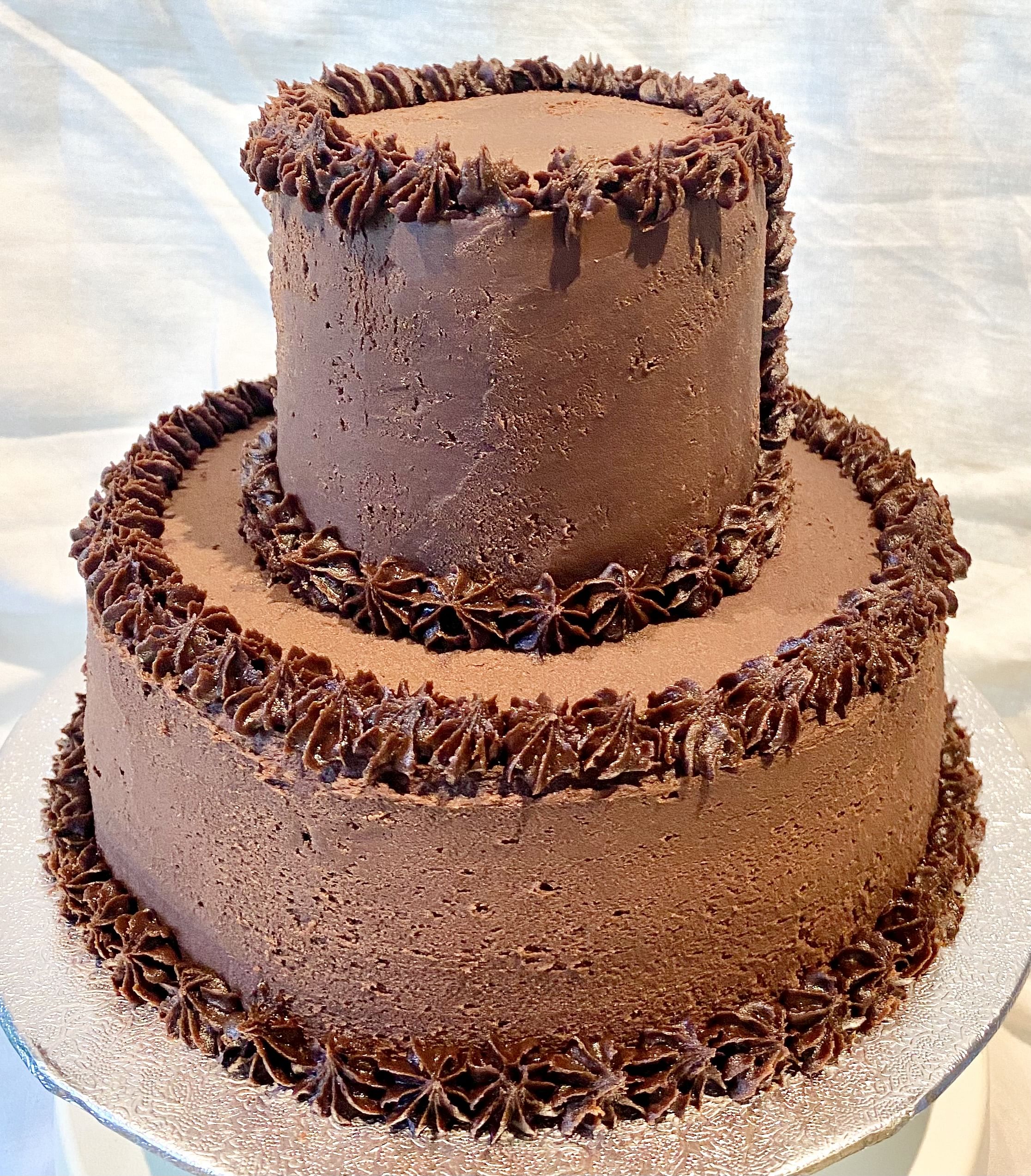 A two-tiered chocolate cake with a smooth, rich chocolate frosting covering both layers. The edges of each tier are decorated with piped chocolate frosting in a star pattern, adding a textured detail to the otherwise smooth surface. The cake sits on a silver, textured cake board, and the background is a soft, light fabric.
