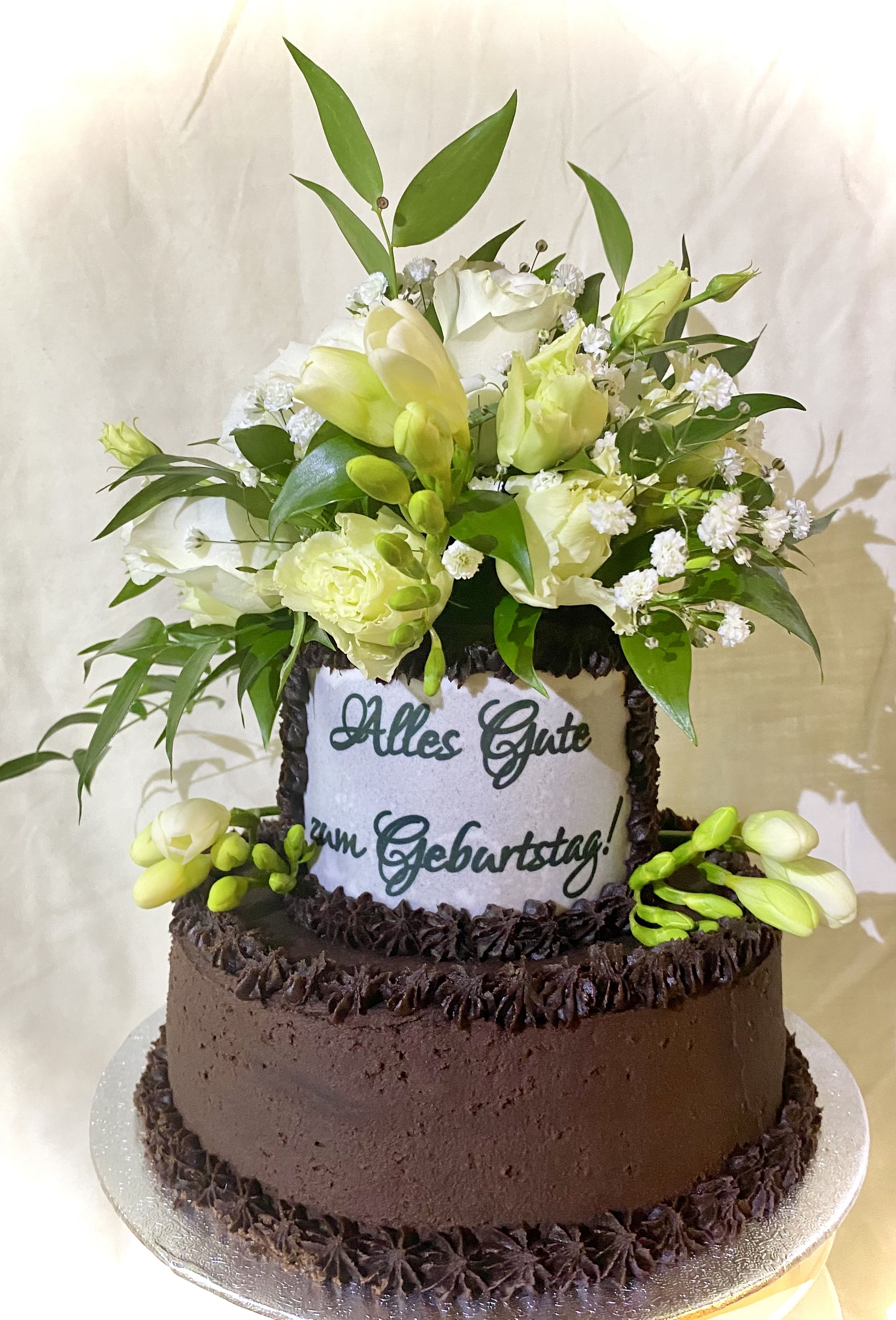 A two-tiered chocolate cake decorated with dark chocolate frosting. The top tier is adorned with a bouquet of white and light green flowers, including roses and other small blossoms, along with green leaves. The cake features a message written in German: 'Alles Gute zum Geburtstag!' which translates to 'Happy Birthday!' The cake is placed on a silver cake board.