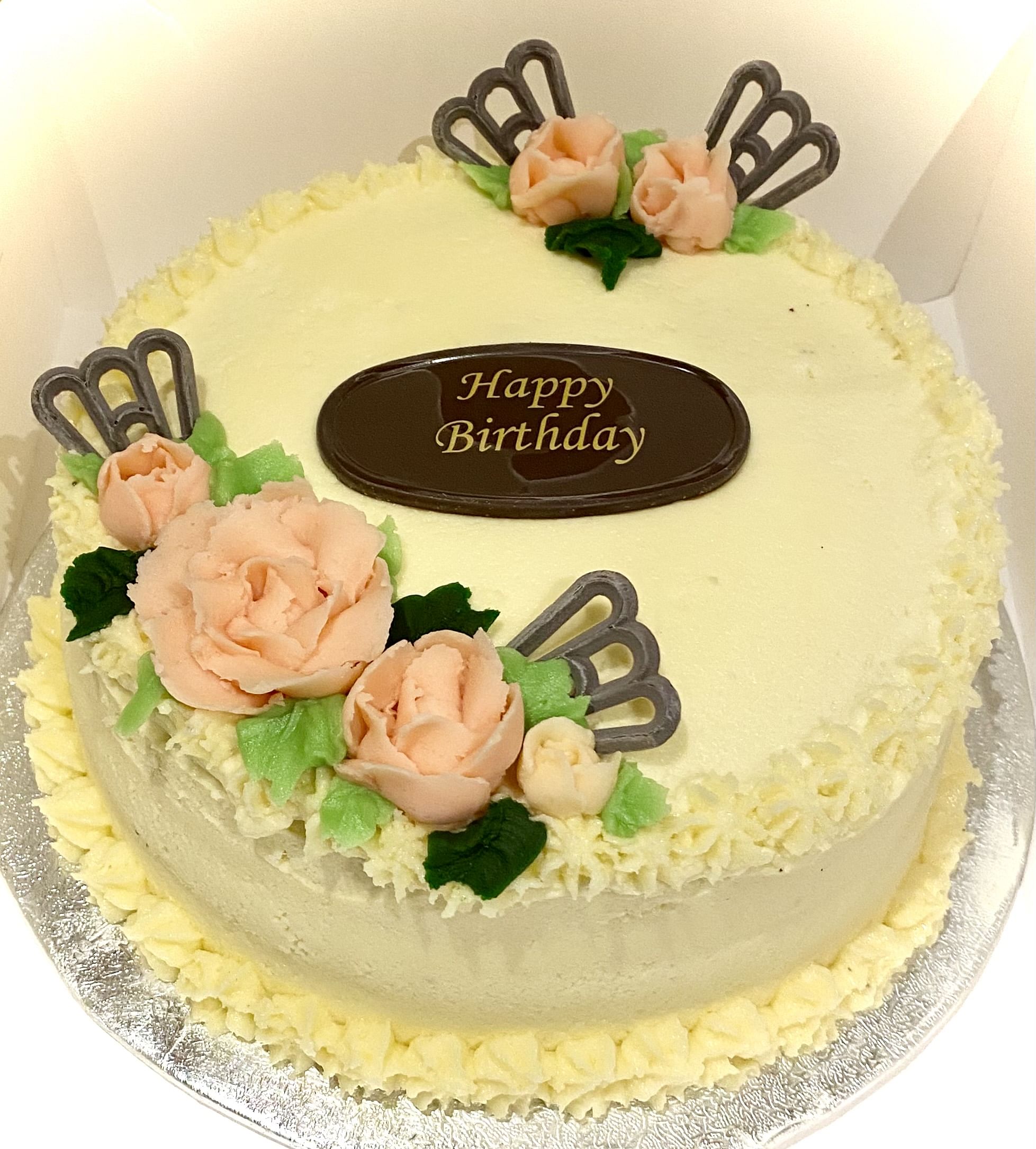 Round birthday cake with smooth, creamy white frosting. Decorated with light pink rose-shaped icing flowers and green leaves. Three decorative chocolate pieces shaped like fans are placed around the flowers. A chocolate plaque in the center reads 'Happy Birthday' in gold lettering. The cake sits on a silver foil-covered base.