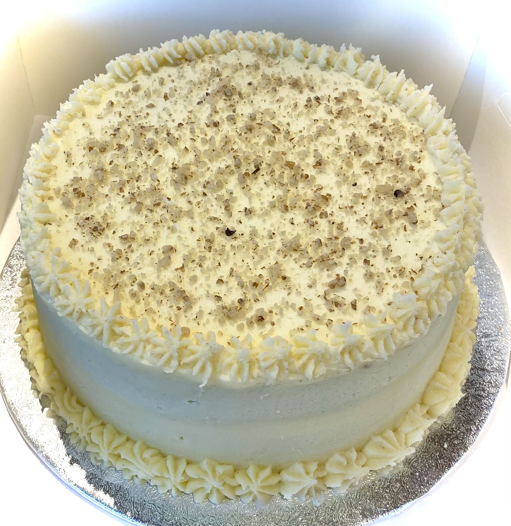 A round cake with a smooth, creamy white frosting covering the sides and top. The top is sprinkled with finely chopped nuts, adding texture and a bit of color contrast. The cake is decorated with a border of piped frosting stars around the top and bottom edges. It is placed on a silver, textured cake board. The cake is inside a white box, which provides a clean background.