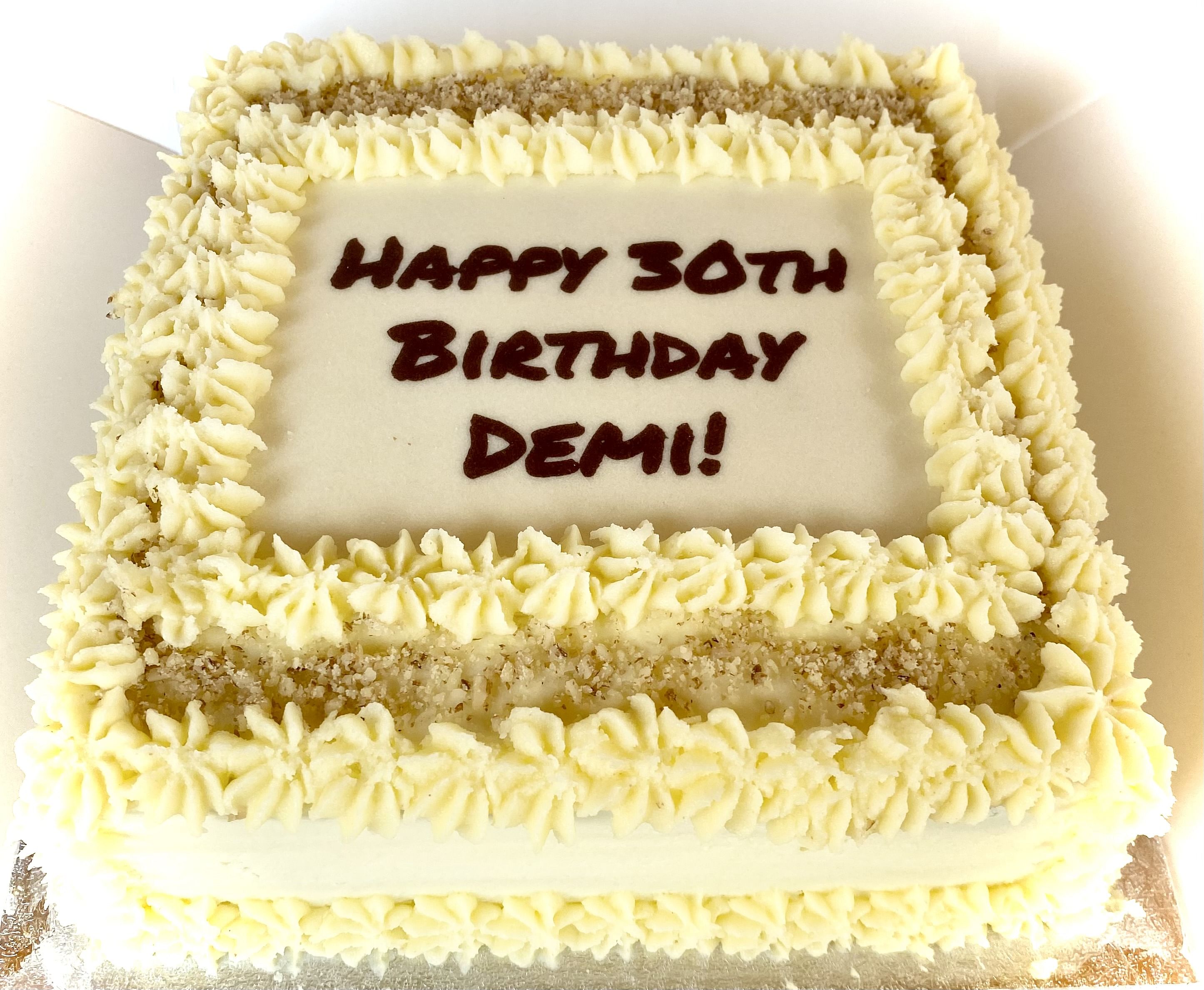 Square cake with white frosting and decorative piped borders. The top has a message written in dark brown icing: 'HAPPY 30TH BIRTHDAY DEMI!' The cake is adorned with small piped rosettes and a sprinkle of crushed nuts around the edges.