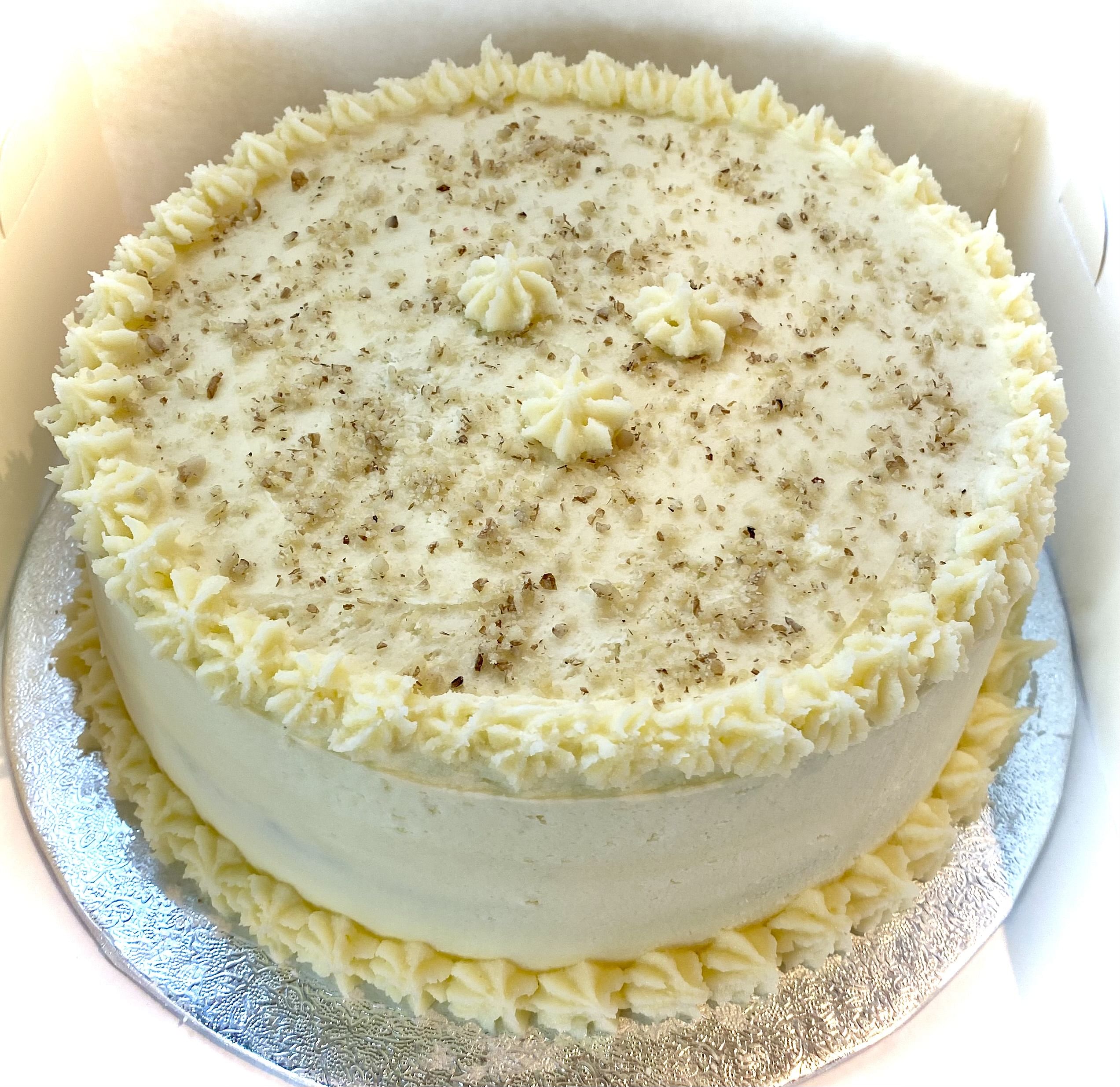 A round cake with a smooth, creamy white frosting. The top is decorated with small piped stars of frosting around the edge and a few in the center. The surface is sprinkled with finely chopped nuts, adding texture and a hint of color. The cake sits on a silver-colored cake board with an embossed pattern.