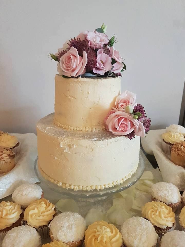 A two-tiered white cake adorned with pink roses and purple flowers on the top and side, displayed on a transparent glass stand. The cake is surrounded by white and yellow frosted cupcakes on a white cloth-covered table.