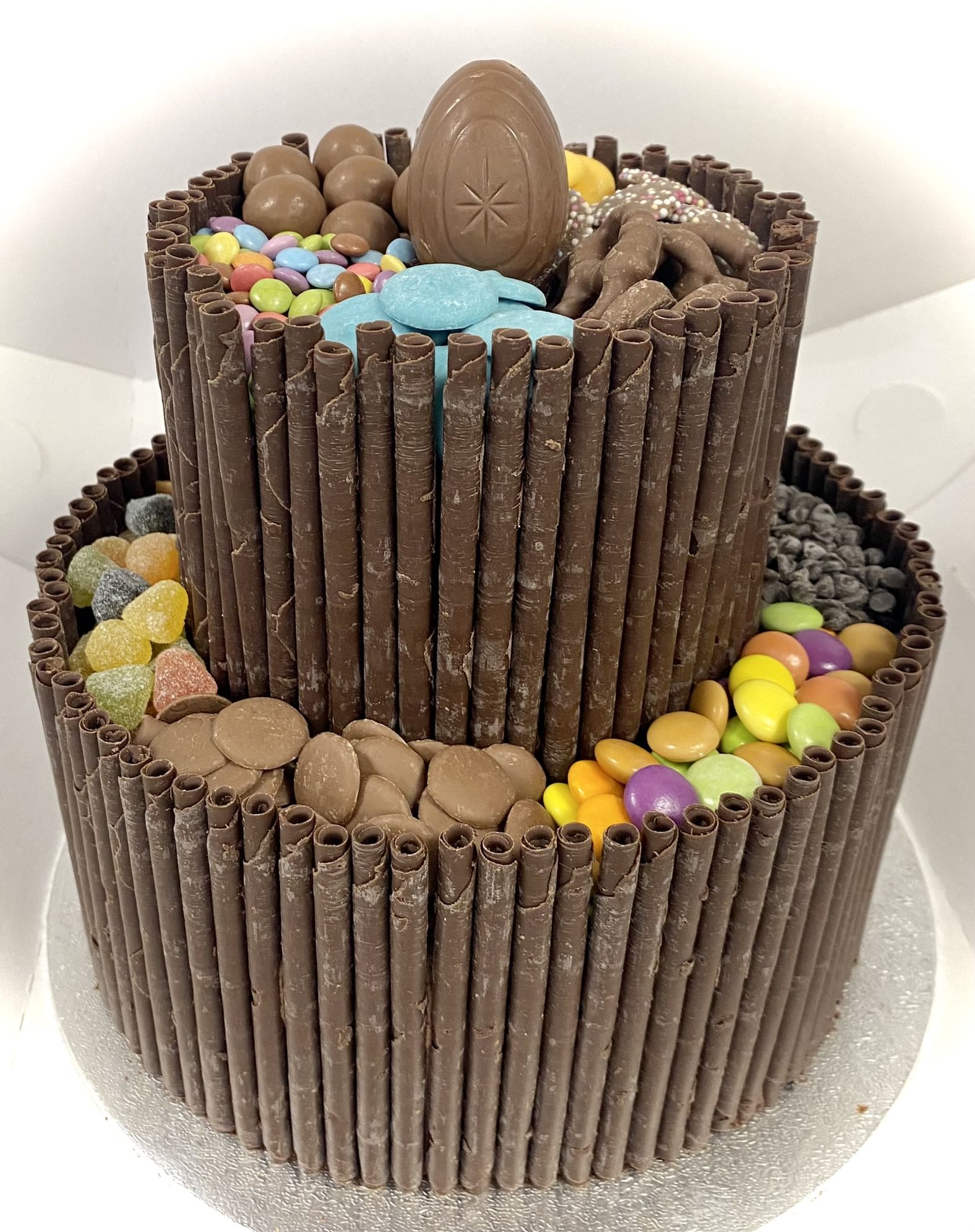 A two-tiered cake adorned with chocolate sticks around the sides. The top tier is decorated with an egg-shaped chocolate and various candies, including gummies and bright-colored chocolates. The bottom tier is similarly decorated with assorted candies and chocolates.