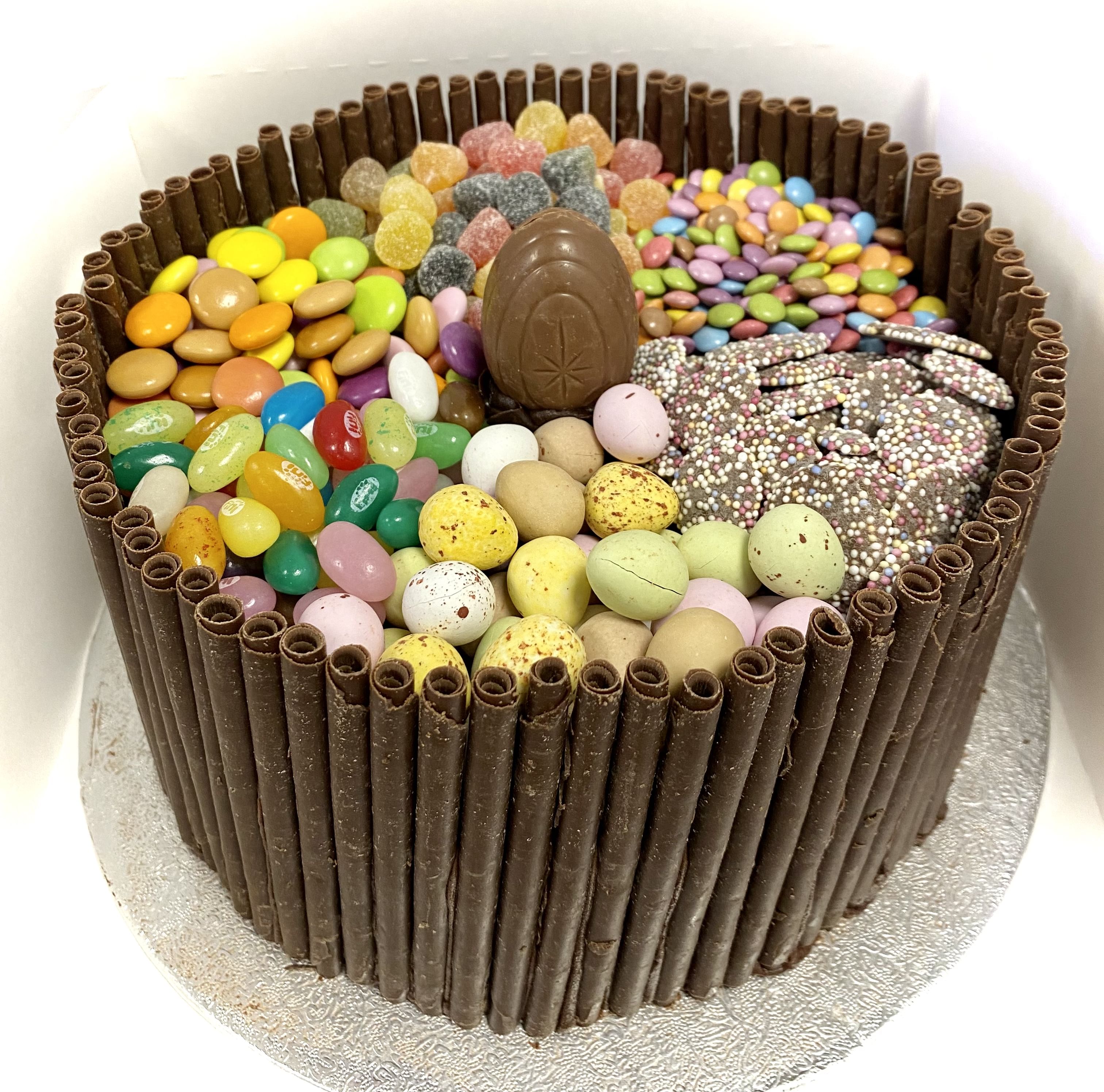 A round cake with a border of vertical chocolate sticks. The top is divided into sections filled with colorful candies including jelly beans, chocolate eggs, gummy bears, and other assorted sweets. The cake sits on a silver board.