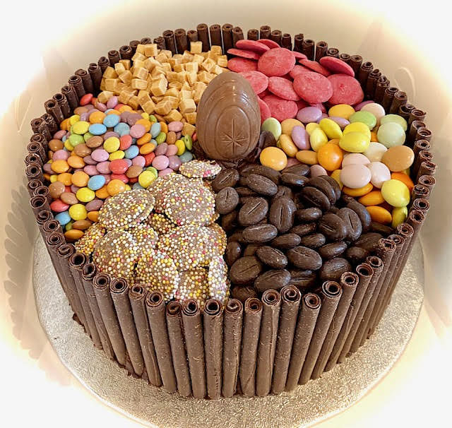 A large round cake is shown, its sides vertically lined with dark chocolate sticks, and the top border is adorned with small chocolate curls. The top of the cake is divided into four distinct sections, each filled with different sweets. Clockwise from the top left, the sections contain: light brown sugar cubes or fudge pieces, flat pink candies, dark brown coffee bean-shaped candies, and brightly colored round candies. In the center of the cake is a standing, elongated chocolate egg with a textured design. Another section of the cake contains flat chocolate disks covered in colorful sprinkles. The cake is presented on a silver cake board within a white box.