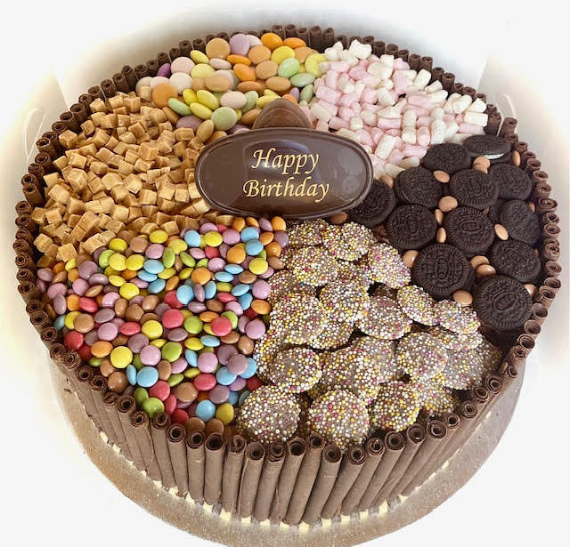 A top-down view shows a round cake with its sides covered in vertical chocolate sticks and a border of chocolate curls. The top of the cake is divided into five sections, each filled with different sweets. These include, clockwise from the top: small pastel-colored candies, light pink and white marshmallows, dark chocolate sandwich cookies (Oreos) along with some brown, possibly chocolate, candy beans, and small colorful round candies. In the fifth section, there are round chocolate disks covered in colorful sprinkles. In the center of the cake, a dark brown oval plaque reads 'Happy Birthday' in a golden script. The cake is on a silver cake board inside a white box.