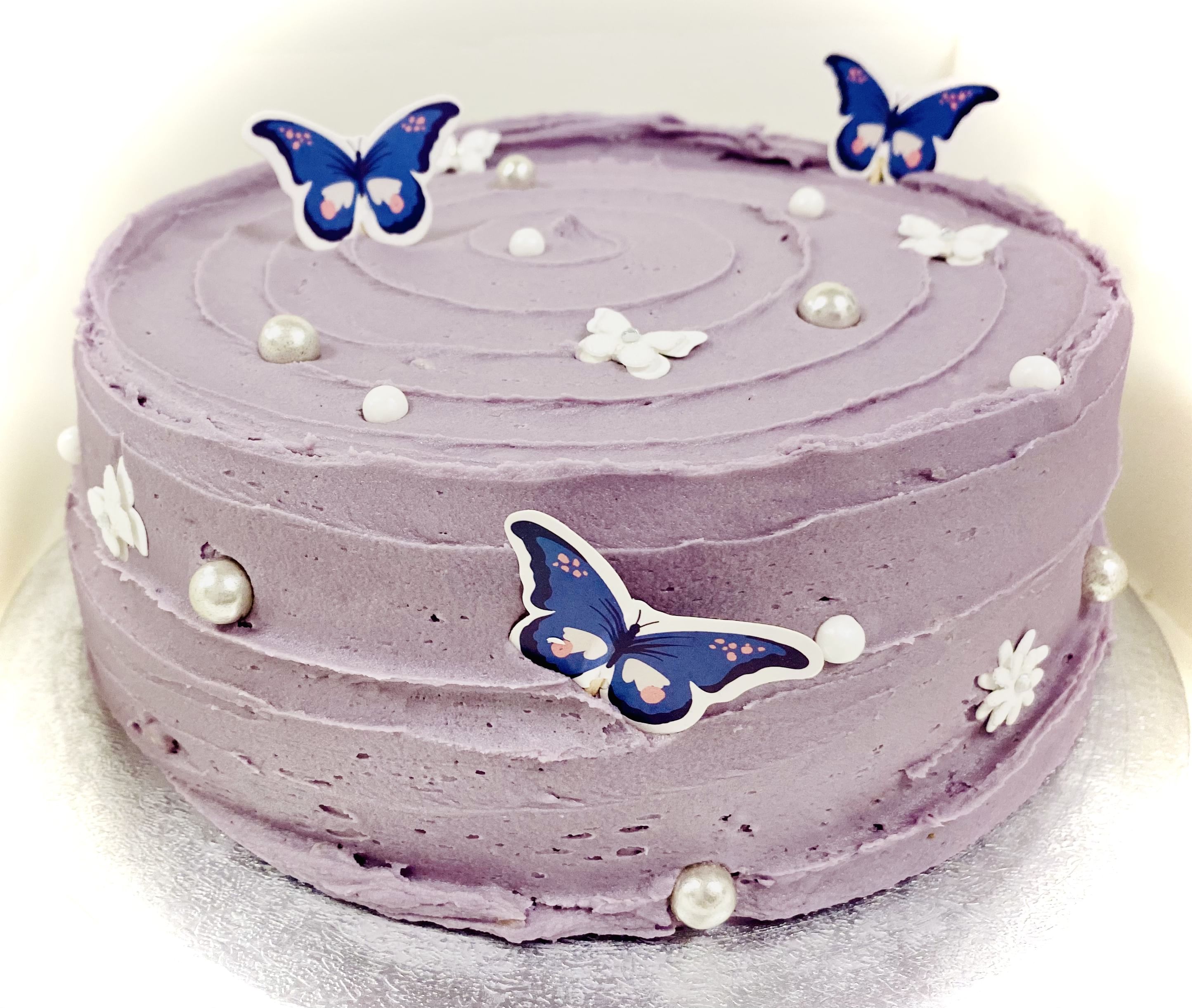 A round, pastel purple custom cake adorned with white pearls, small white flower decorations, and blue butterfly toppers. The frosting is textured in a spiral pattern. This custom dessert is placed on a silver cake board, perfect for an unforgettable event.