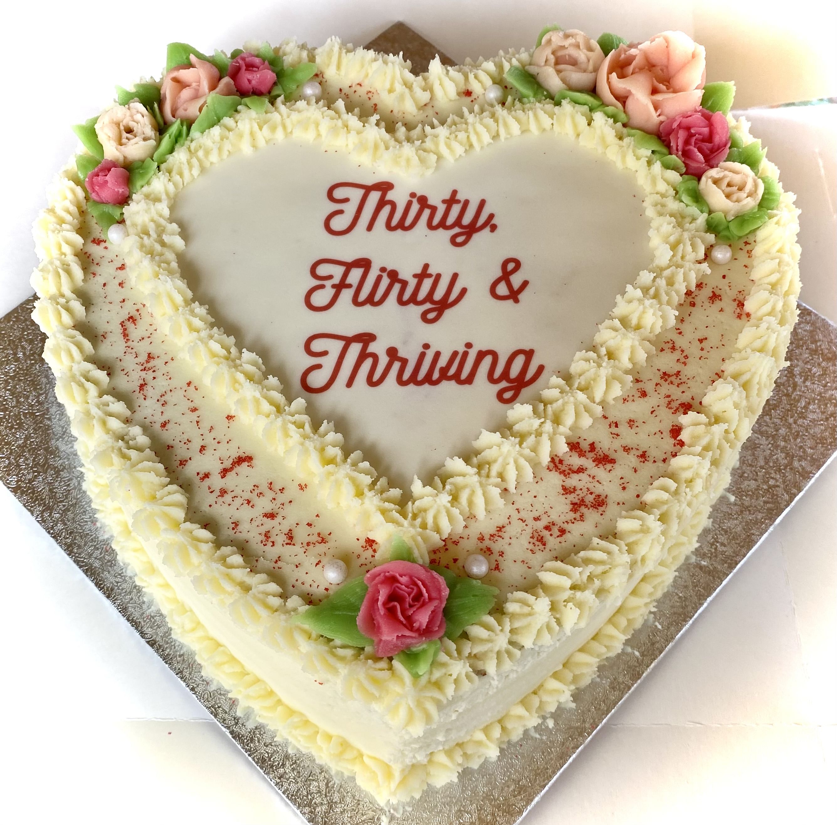 Heart-shaped cake with white frosting and decorative cream flowers in shades of pink and peach. The cake is bordered with piped cream and sprinkled with red specks. In the center, the text reads: 'Thirty, Flirty & Thriving' in red cursive font. The cake sits on a silver board.
