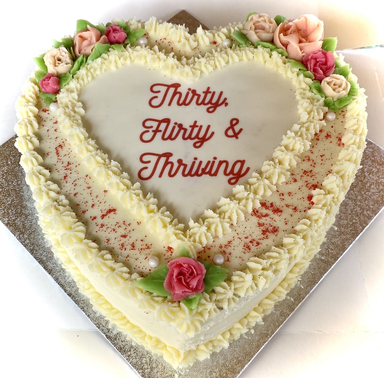 Heart-shaped cake with white frosting and decorative cream flowers in shades of pink and peach. The cake is bordered with piped cream and sprinkled with red specks. In the center, the text reads: 'Thirty, Flirty & Thriving' in red cursive font. The cake sits on a silver board.