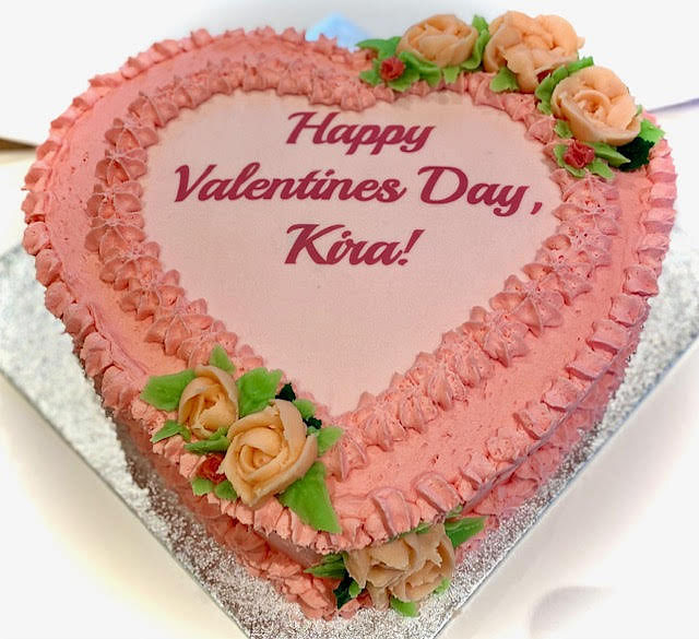 Heart-shaped cake with pink frosting and decorative peach-colored roses with green leaves. The cake has a message written on top in red lettering: 'Happy Valentines Day, Kira!' It is placed on a silver-colored board.