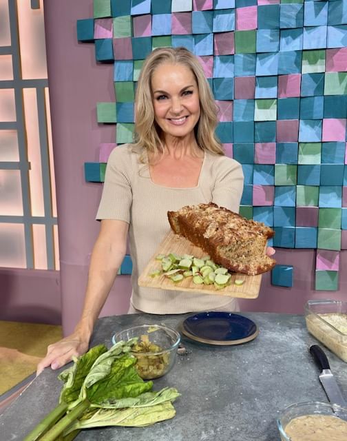 A smiling woman with blonde hair, wearing a beige short-sleeved top, holds up a loaf of freshly baked bread on a wooden cutting board. The board also has slices of chopped rhubarb. In the foreground, there are rhubarb stalks, a glass bowl of chopped fruit, and ingredients for baking on a counter. The background is a brightly colored set with a wall of stacked, multi-colored cubes.