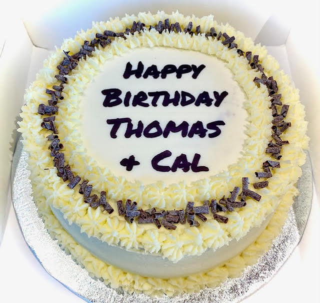A round cake, likely a birthday cake, is frosted with pale yellow icing and features a central white circular area. Around the outer edge of the cake, there is a border of piped yellow frosting with dark chocolate shavings scattered on top. The smooth white center of the cake is inscribed with 'HAPPY BIRTHDAY THOMAS + CAL' in dark purple lettering. The cake is presented on a silver cake board within a white box.