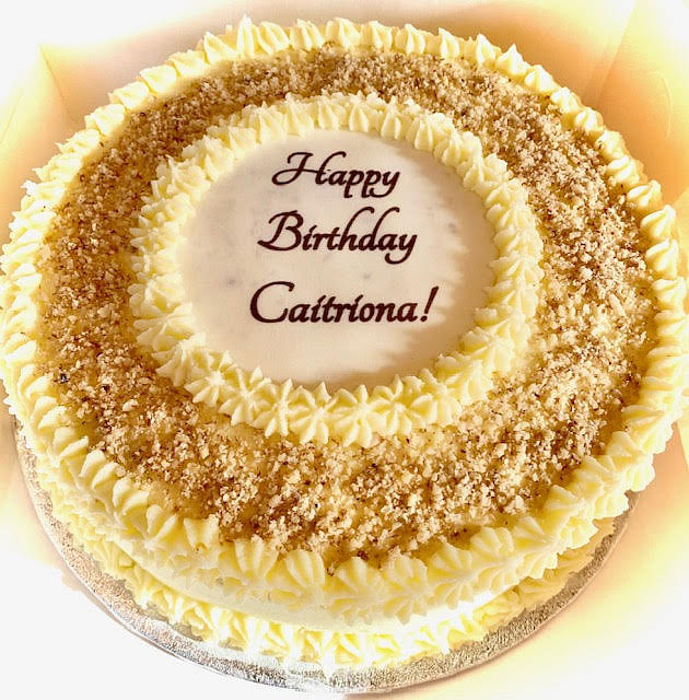 A round white cake is shown, with 'Happy Birthday Catriona!' written in dark script in the center. The cake is decorated with concentric rings: an inner ring of white frosting, followed by a ring of light brown crumbs or nuts, then a ring of white frosting rosettes, and finally an outer ring of light brown crumbs or nuts, bordered by more white frosting rosettes. The cake rests on a silver cake board.