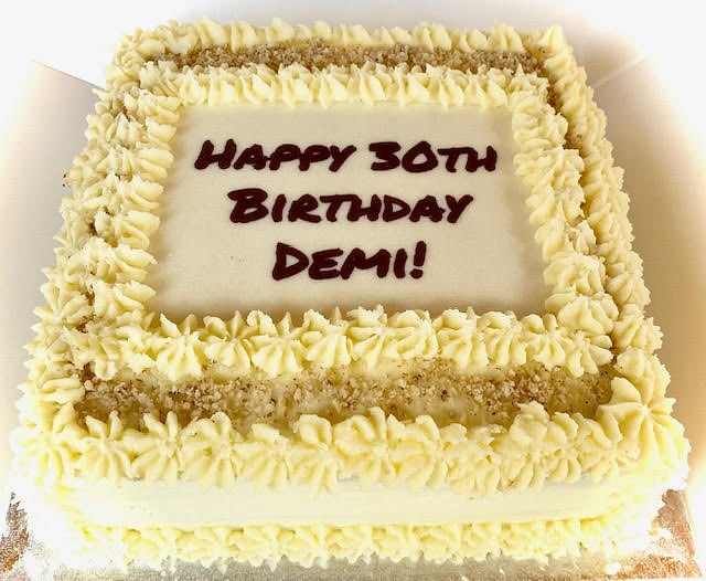 A square cake, possibly a lemon wonder cake, is frosted with pale yellow icing and decorated with piped swirls around the edges and a central rectangular area. The piped swirls create a decorative border, and within this border, there are two horizontal lines of light brown crumble or nuts. The smooth, white rectangular area in the center of the cake is inscribed with 'HAPPY 30TH BIRTHDAY DEMI!' in dark brown lettering. The cake rests on a silver cake board within a white box.