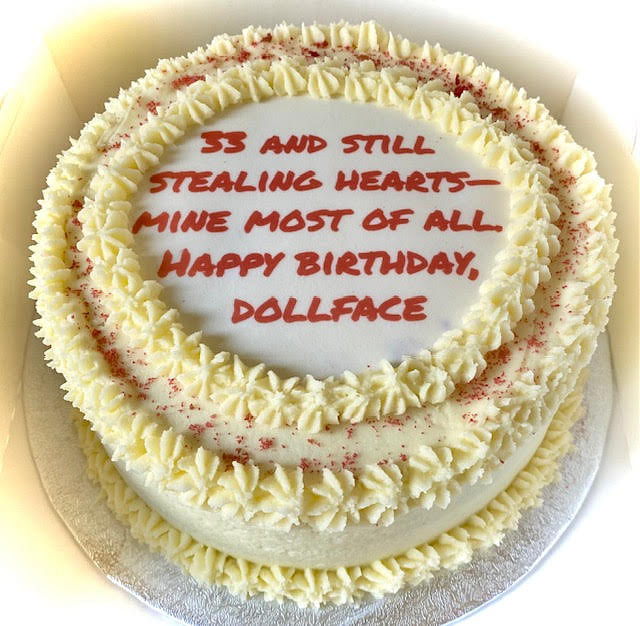 A round white cake is topped with red frosting that reads: '33 AND STILL STEALING HEARTS— MINE MOST OF ALL. HAPPY BIRTHDAY, DOLLFACE'. The cake has multiple concentric rings of piped white frosting rosettes and is dusted with red sprinkles. It is presented on a silver cake board.