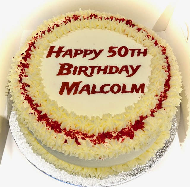 A round white cake with 'Happy 50th Birthday Malcolm' written in red frosting in the center. The cake is decorated with a border of white frosting rosettes and sprinkled with red crumbs or sprinkles. It sits on a silver cake board.