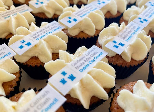 A close-up shot of several cupcakes with light yellow frosting, each topped with a white rectangular edible decoration featuring the text 'Comhairle Cathrach Bhaile Átha Cliath Dublin City Council' and a blue logo of three castles.