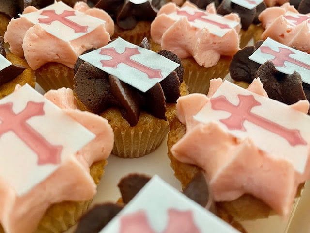 A close-up shot shows several cupcakes with pale pink and chocolate frosting, each topped with a white square edible decoration featuring a pink cross.