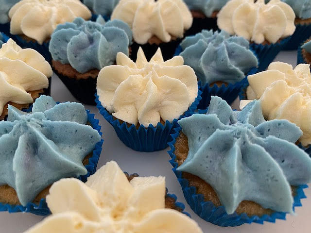 A close-up shot shows an assortment of cupcakes with piped frosting. Some cupcakes have light blue frosting, and others have cream-colored frosting, both swirled into a star or rosette shape. They are all in blue cupcake liners. The background is slightly blurred.
