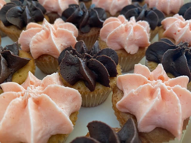 A close-up shot of several mini cupcakes, some topped with light pink frosting piped in a star shape, and others with dark brown chocolate frosting also piped in a star shape. The cupcakes are in light-colored paper liners and arranged closely together.