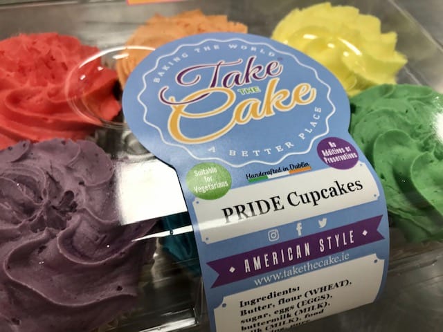 An assortment of pride cupcakes in vibrant colors, including red, orange, yellow, green, blue, and purple, arranged in a plastic container. The label on the container reads 'Pride Cupcakes' and includes references to 'American Style' and the bakery's website and ingredients.