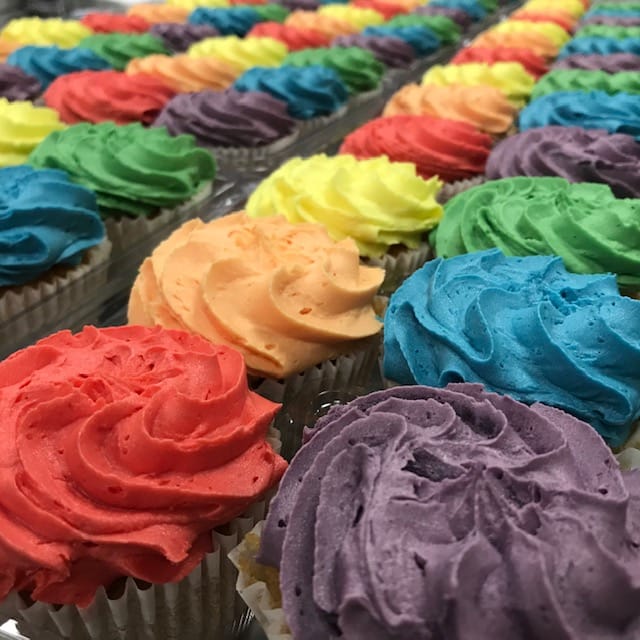 A close-up image of colorful Pride Cupcakes arranged in rows. The cupcakes are topped with swirls of frosting in various vibrant colors, including red, orange, yellow, green, blue, and purple, creating a visually striking and appetizing display.