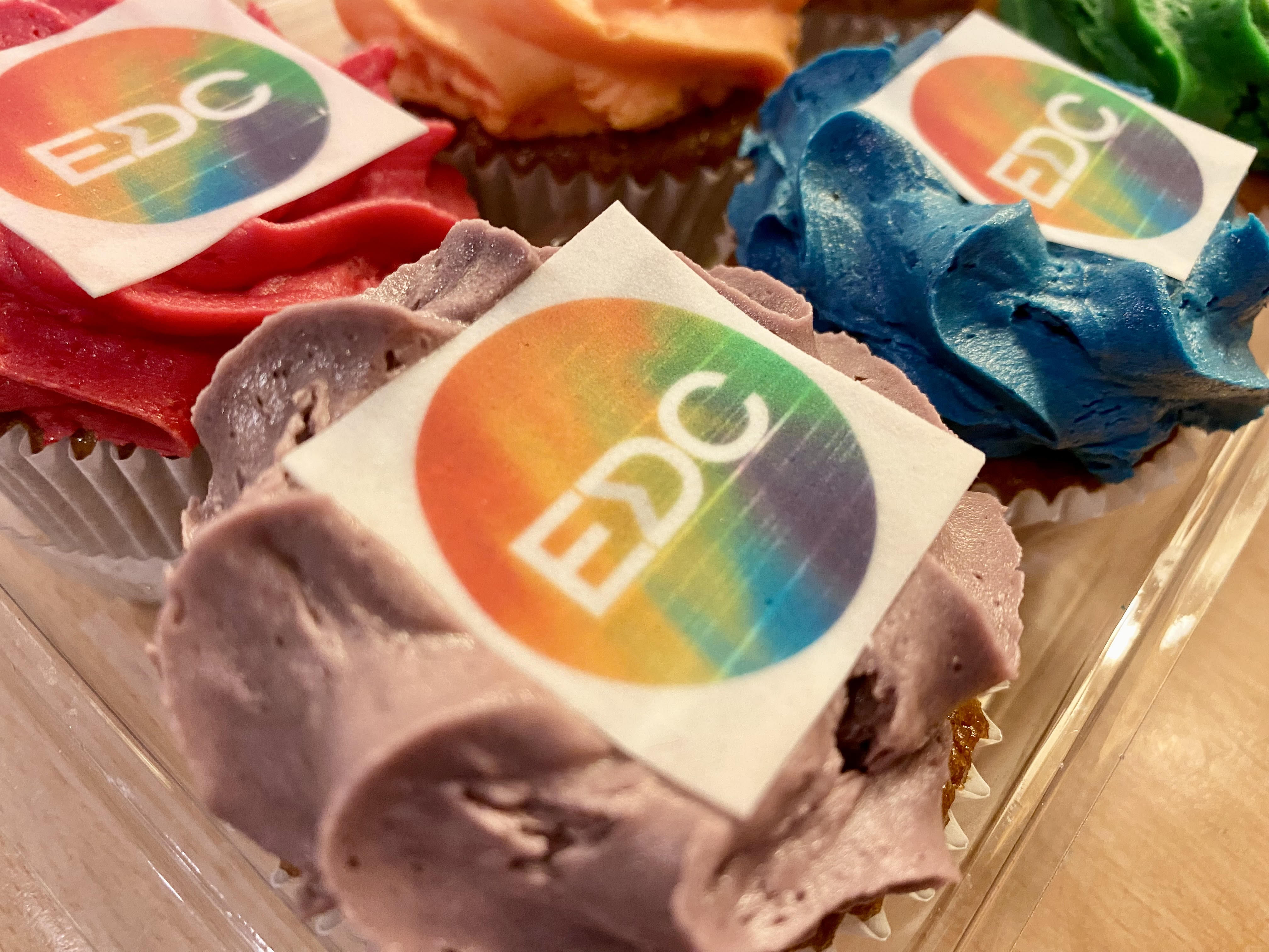 A close-up of cupcakes with brightly colored frosting, including red, blue, and purple. The cupcakes are decorated with an edible square topper that has a circular rainbow design and the letters 'EDC'.