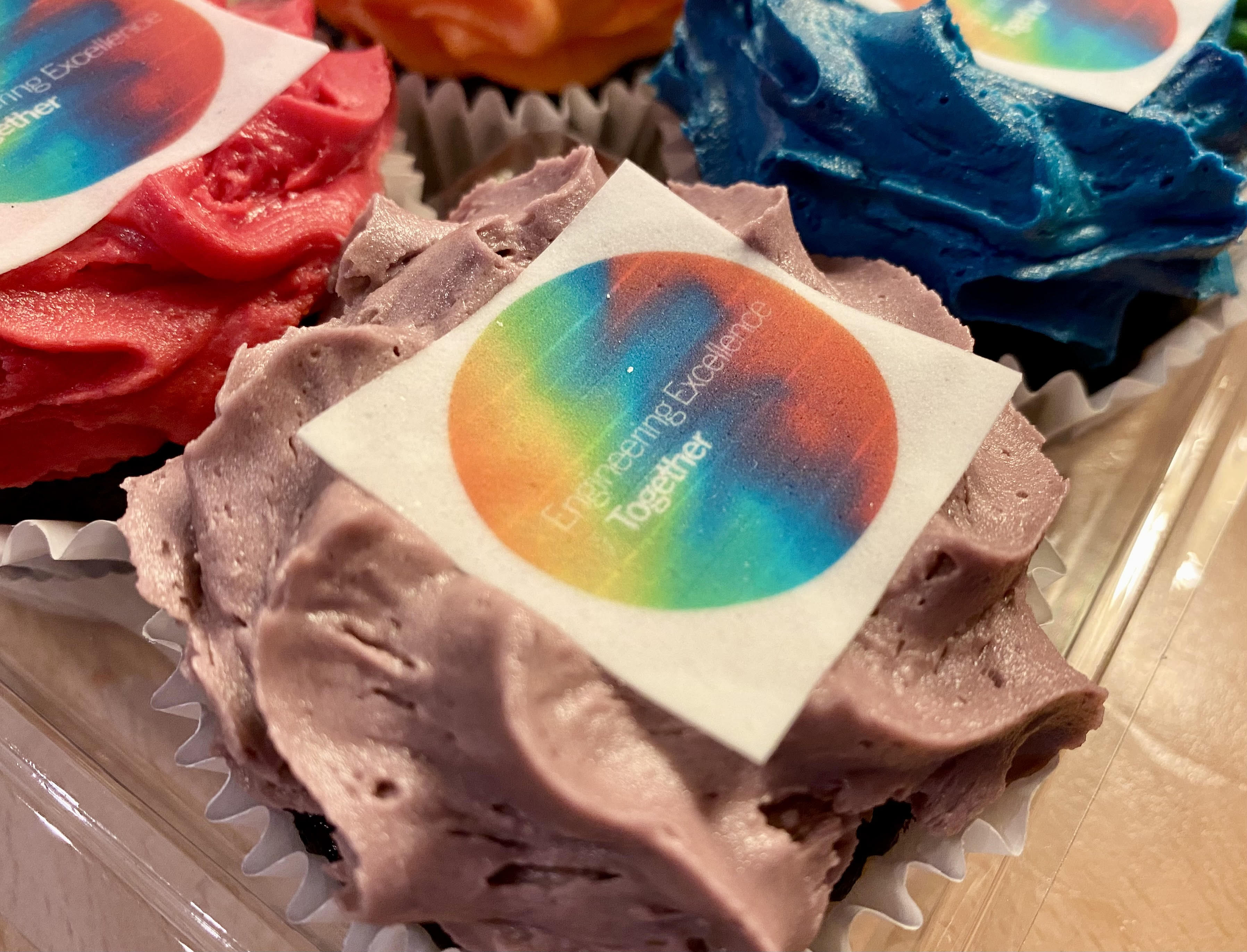 A close-up of cupcakes with colorful frosting in shades of red, blue, and purple. On top of the frosting is an edible square topper with a circular rainbow design and the words 'Engineering Excellence Together'.