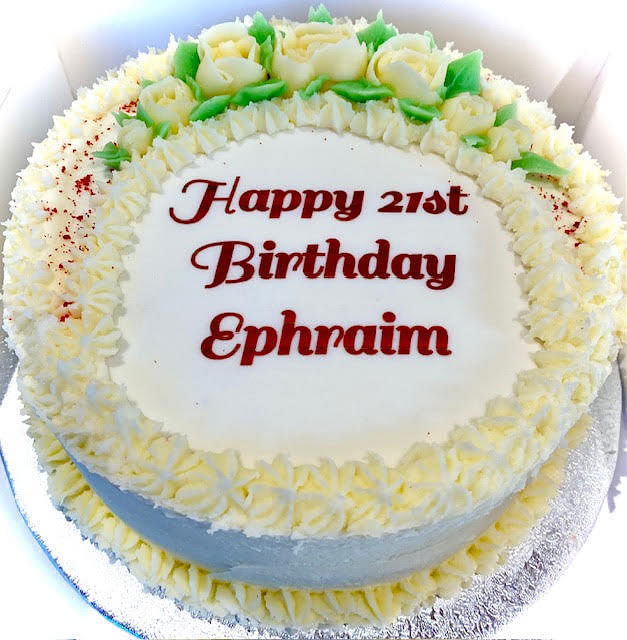 A round white cake is adorned with 'Happy 21st Birthday Ephraim' written in red script in the center. The top edge of the cake is decorated with white frosting roses and green leaves, with red sprinkles dusted over the white frosting rosettes that frame the top of the cake. The cake rests on a silver cake board.