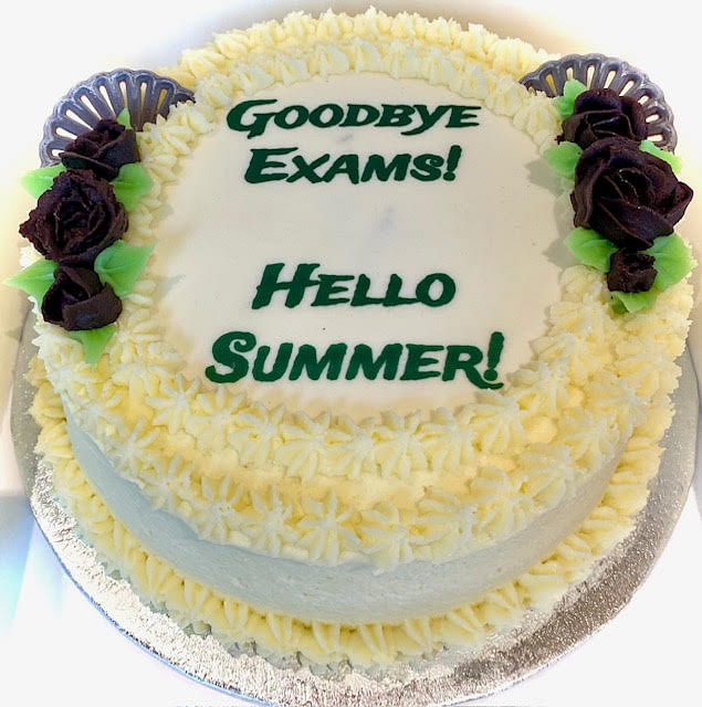 A round white cake is topped with the phrases 'GOODBYE EXAMS!' and 'HELLO SUMMER!' written in green frosting. The cake is decorated with a border of yellow-tinted white frosting rosettes, and features dark purple or black frosting roses with green leaves, and two dark fan-shaped decorations on opposite sides of the top. The cake is on a silver cake board.