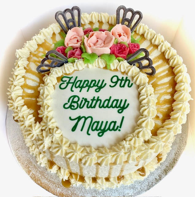 A round white cake is inscribed with 'Happy 9th Birthday Maya!' in green cursive font in the center. The cake is decorated with a border of white frosting rosettes and caramel drizzles over the top. It is also adorned with pink and red frosting roses with green leaves, and two dark, butterfly-like or fan-shaped chocolate decorations on either side of the roses. The cake is presented on a silver cake board.
