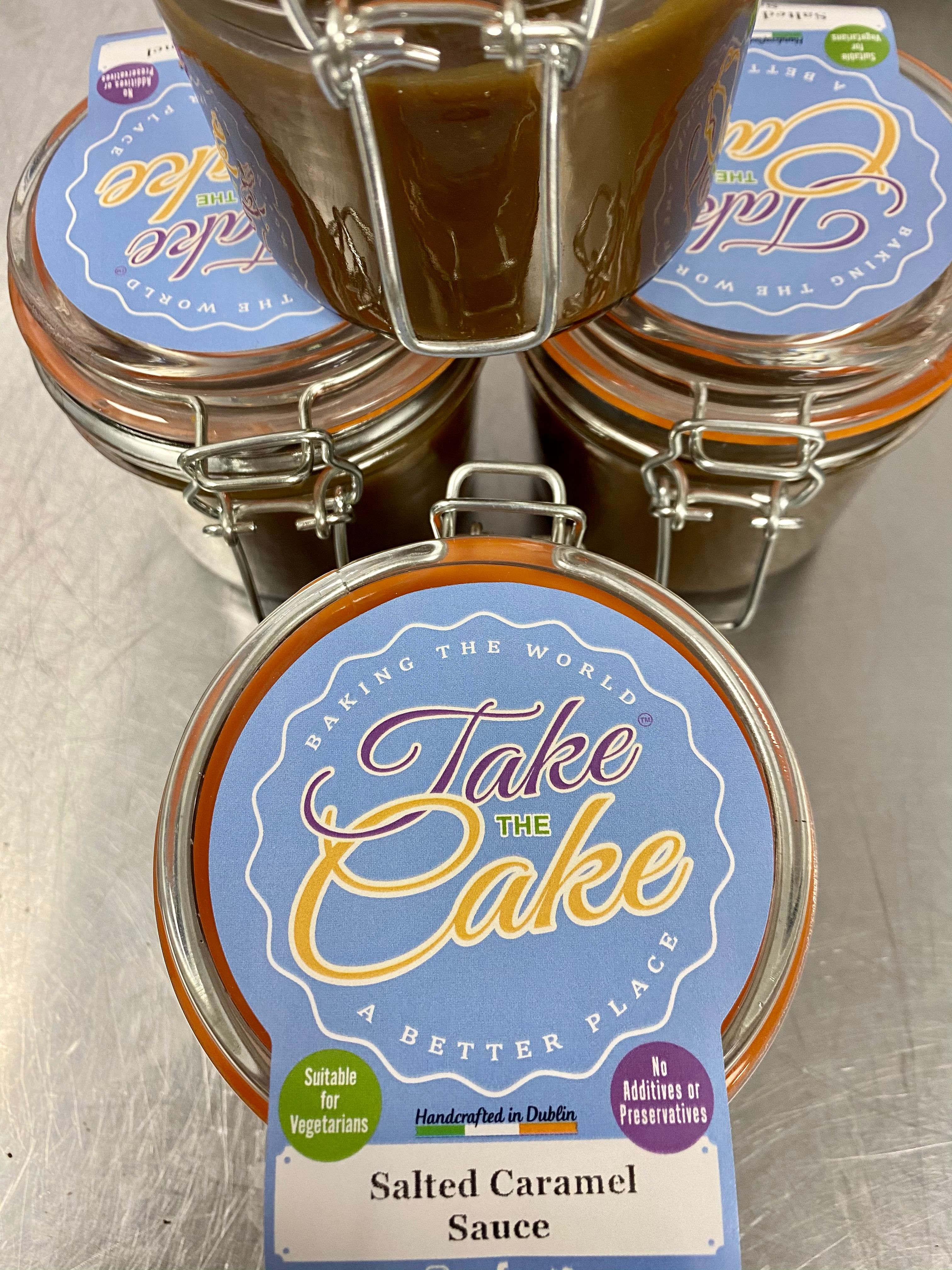 Three glass jars filled with salted caramel sauce are shown on a stainless steel surface. The jars have blue lids with a label that reads 'Take the Cake' and other details including 'Baking the World a Better Place,' 'Suitable for Vegetarians,' 'Handcrafted in Dublin,' and 'No Additives or Preservatives'.