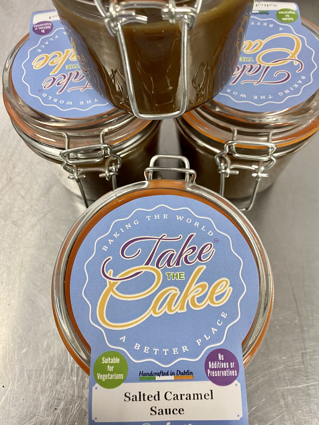 Three glass jars filled with salted caramel sauce are shown on a stainless steel surface. The jars have blue lids with a label that reads 'Take the Cake' and other details including 'Baking the World a Better Place,' 'Suitable for Vegetarians,' 'Handcrafted in Dublin,' and 'No Additives or Preservatives'.