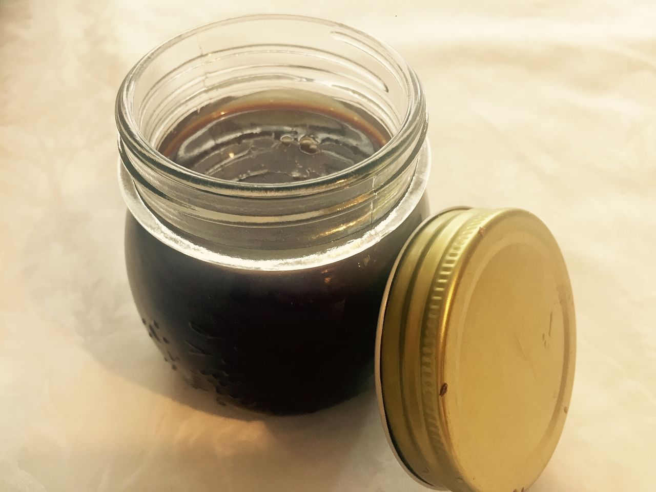A glass jar filled with a dark liquid, possibly salted caramel sauce or syrup, with a gold-colored metal lid placed beside the jar on a light surface. The jar has measurement markings and is illuminated by a soft light, embodying homemade goodness.