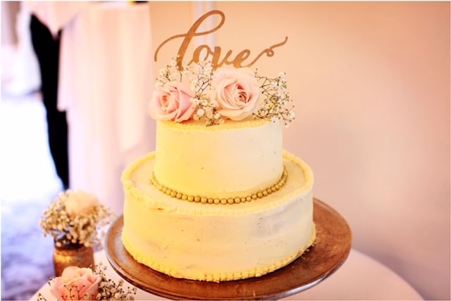 A two-tiered white cake stands on a wooden cake stand. The top tier is adorned with pink roses and small white flowers, accompanied by a decorative gold 'Love' topper. Ideal for special occasions, this perfect dessert complements the soft, warmly lit environment, creating an unforgettable event.