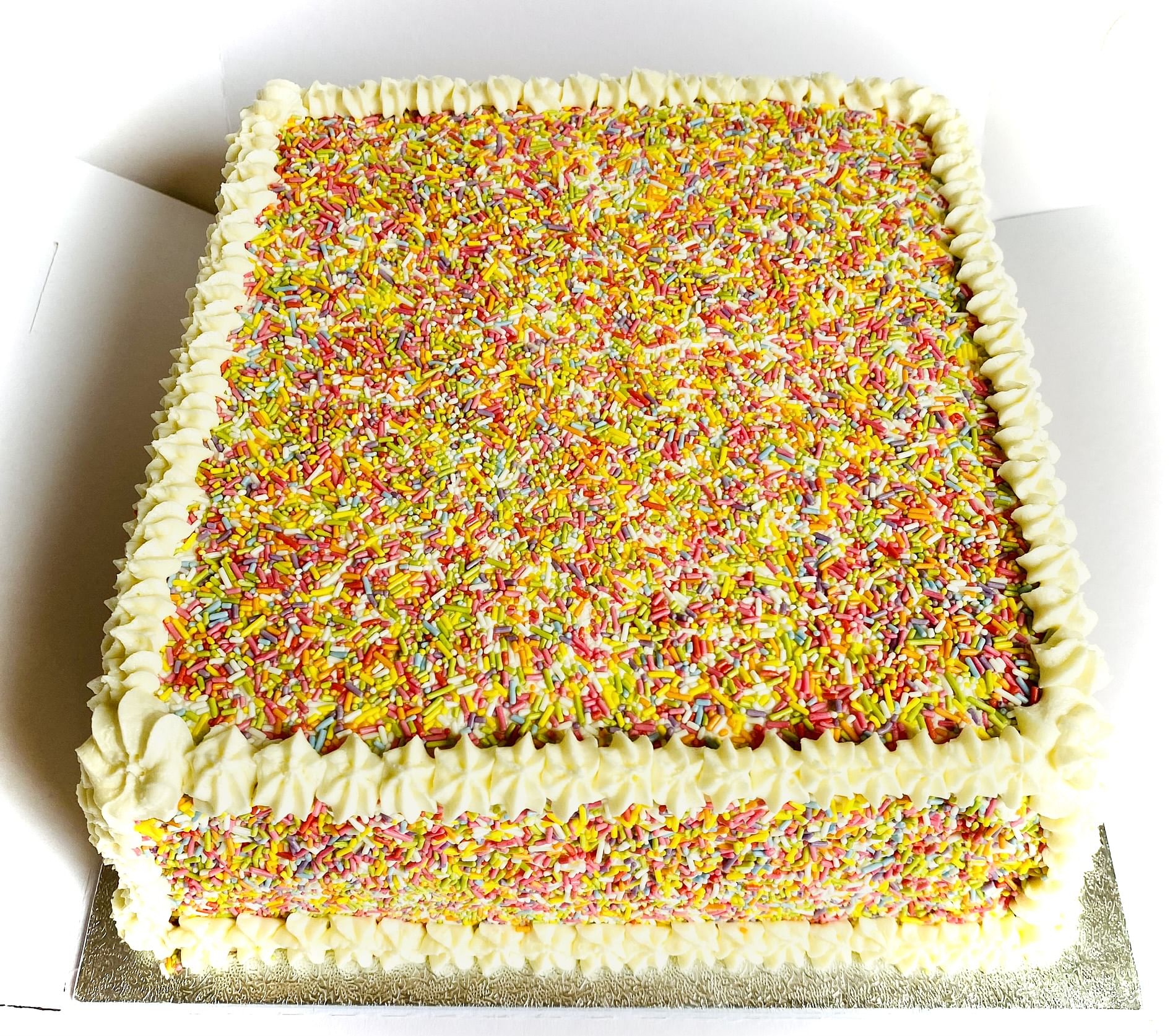 A square sprinkles cake covered in white frosting and decorated with multicolored sprinkles on the top and sides. The frosting is piped in a ruffled pattern along the edges, making it a festive and vibrant celebration centerpiece perfect for birthday parties.