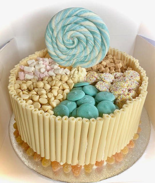 A round cake is featured, its sides decorated with upright white chocolate sticks. The top of the cake is segmented and filled with various confectioneries: pale yellow chocolate chips, small pink and white marshmallows, light blue disc-shaped candies, and circular white candies adorned with colorful sprinkles. A large, flat, swirled blue and white lollipop stands at the back of the cake. The base of the cake is edged with small, clear, amber-colored candies. The cake is presented on a silver cake board within a white box.