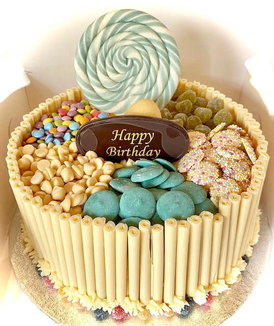 A colorful birthday cake decorated with various candies and a large lollipop. The sides of the round cake are covered with vertical white chocolate sticks. The top of the cake is divided into sections, each filled with different sweets: pale yellow chocolate chips, blue disc-shaped candies, small colorful coated candies, and what appears to be pink and white sprinkles or small candies. A large, swirled green and white lollipop stands upright in the center back, behind a dark chocolate oval plaque that reads 'Happy Birthday' in gold script.